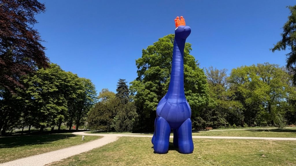 Een grote blauwe opblaasdino staat op een grasveld in een park met bomen en een zandpad.
